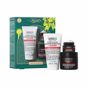 A graphic featuring a dark teal Kiehl’s gift box with white and gold accents on the left. The box contains three products. To the right of the box, the same three products are displayed outside of the box: a white tube of "KIEHL’S ULTRA FACIAL BARRIER-HYDRATING CLEANSER," a small black and red jar of "KIEHL’S AGE DEFENDER EYE REPAIR," and a larger black and red jar of "KIEHL’S AGE DEFENDER CREAM MOISTURIZER." The box is decorated with bright green graphic paint splatters and a bow design and features the text "YOUTH STILL GOT IT" at the bottom.