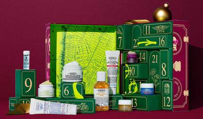 Kiehl’s Advent Calendar gift box in deep red with gold text, displayed next to 24 mini skincare products including Ultra Facial Cream, Calendula Toner, Retinol Serum, and Lip Balm #1. A pink spray-paint graphic on the side calls out the value of the set.
