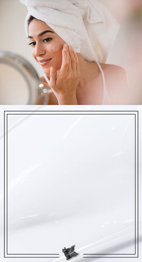 A person with a bath towel on their head, massaging a cream into their face.