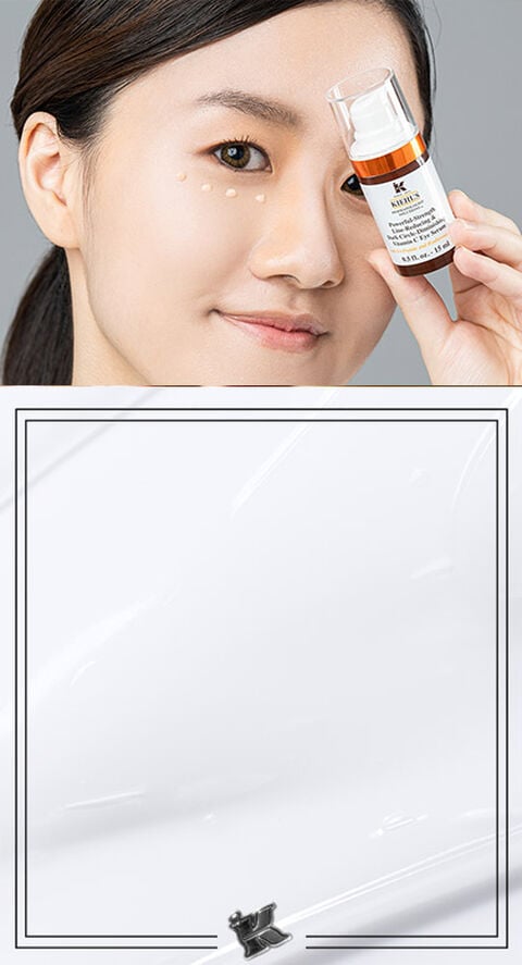 A woman holding Kiehl's Powerful Vitamin C Eye Serum with drops of cream under her eyes.